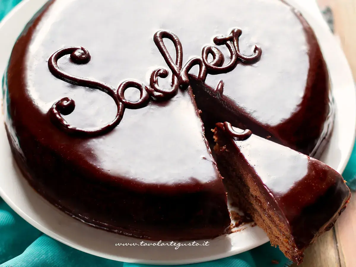 Una lucida torta Sacher, con una fetta tagliata, mostra la sua ricca copertura al cioccolato e la scritta "Sacher" decorata.