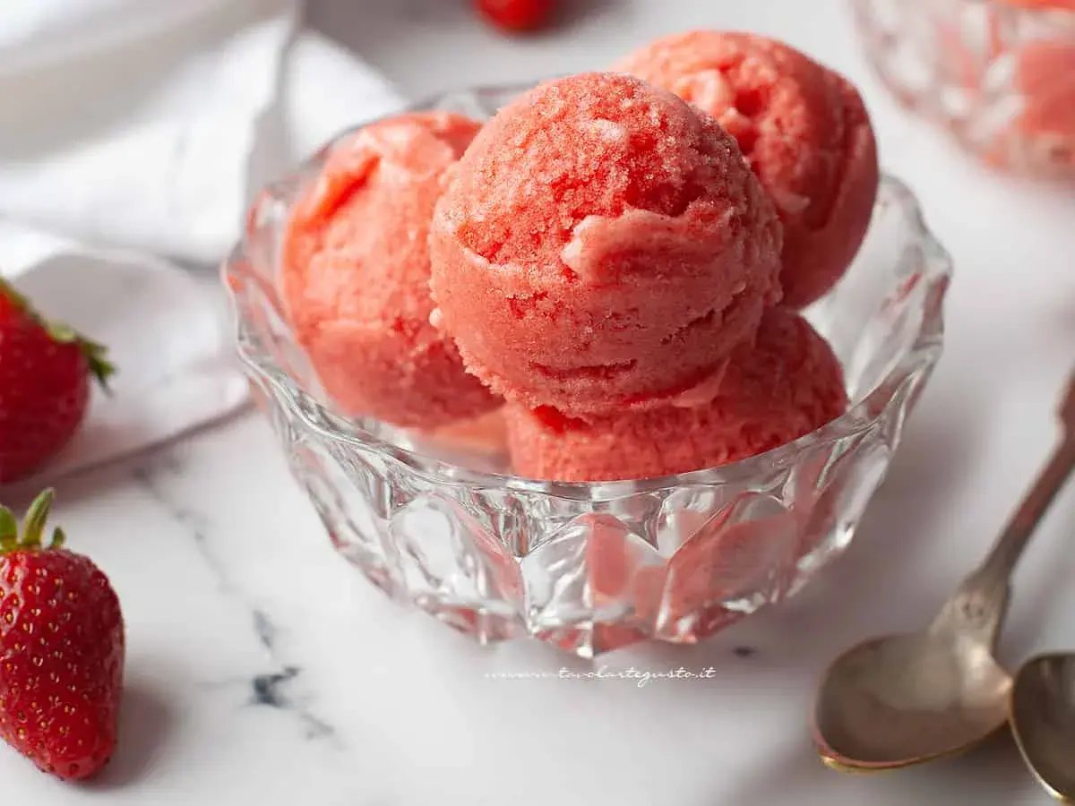 Coppa di sorbetto cremoso alle fragole, fresca e invitante, con fragole fresche e cucchiaini d'argento su sfondo marmorizzato.