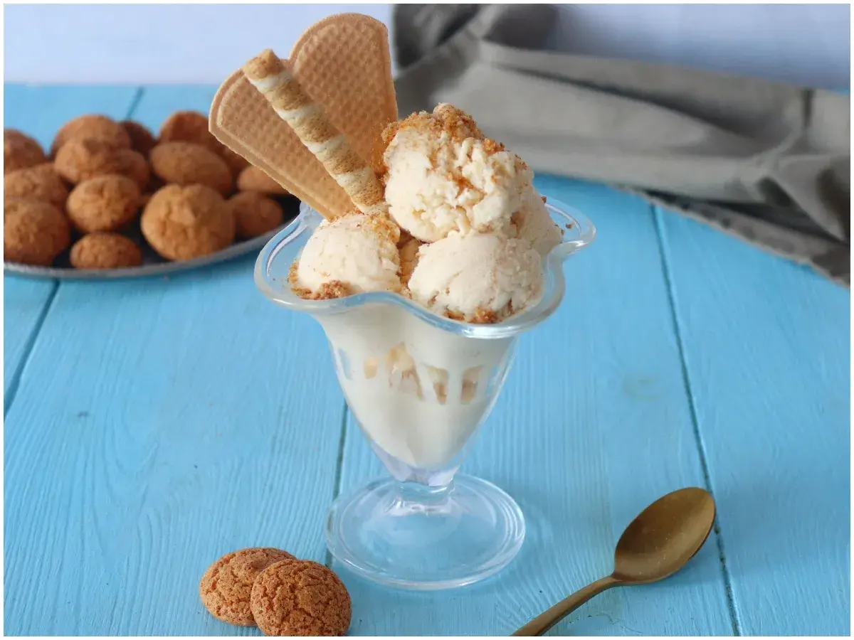 Coppa di gelato amaretto con biscotti e cialde su tavolo blu.