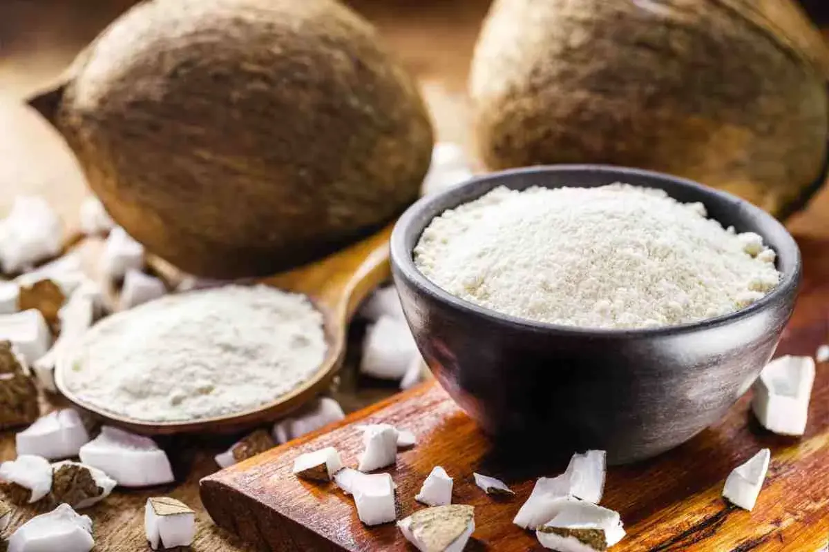 Cocco grattugiato in una ciotola e un cucchiaio, con noci di cocco intere e pezzi di cocco sullo sfondo. Ideale per sostituire la farina di cocco.