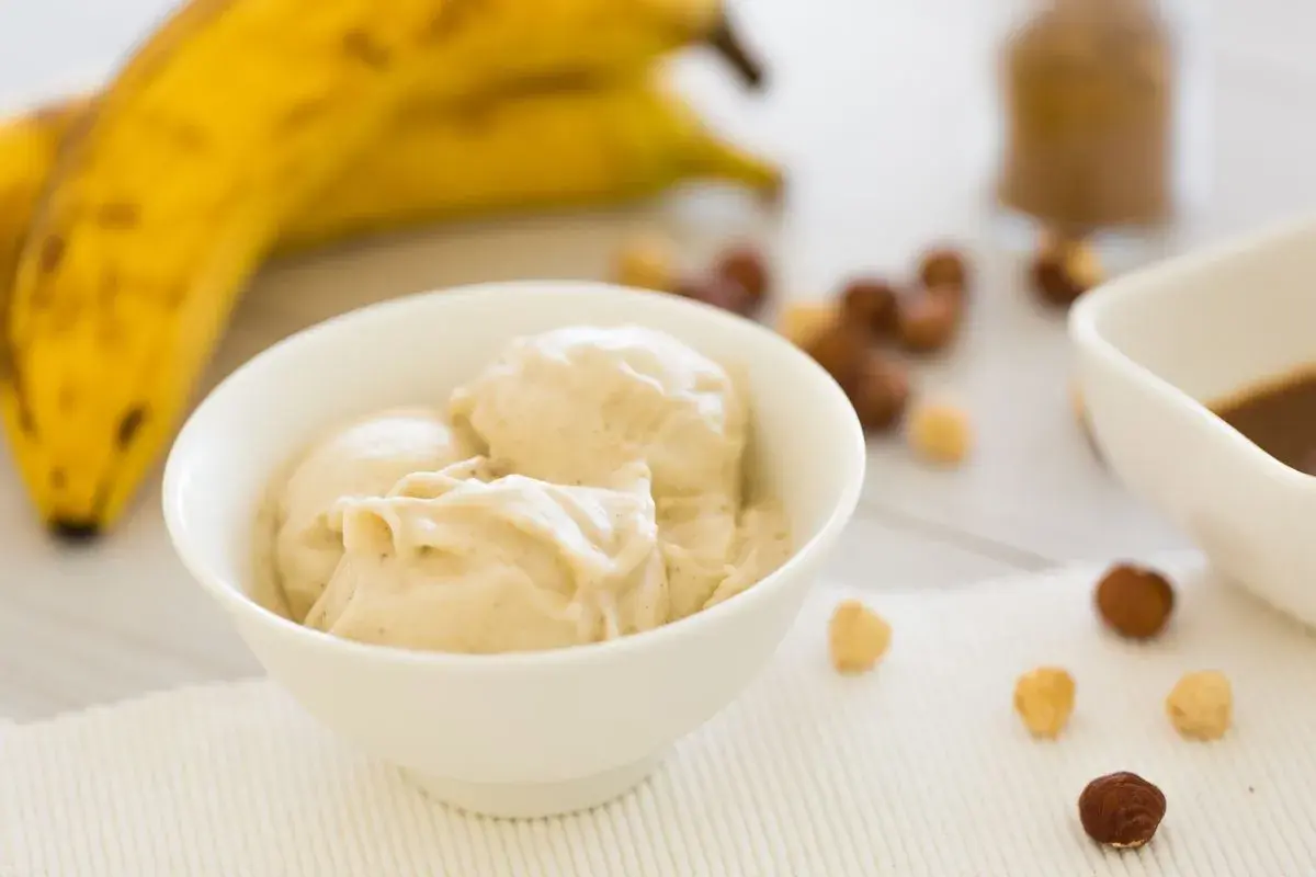 Una ciotola di cremoso gelato alla banana, con banane mature sullo sfondo e nocciole sparse.