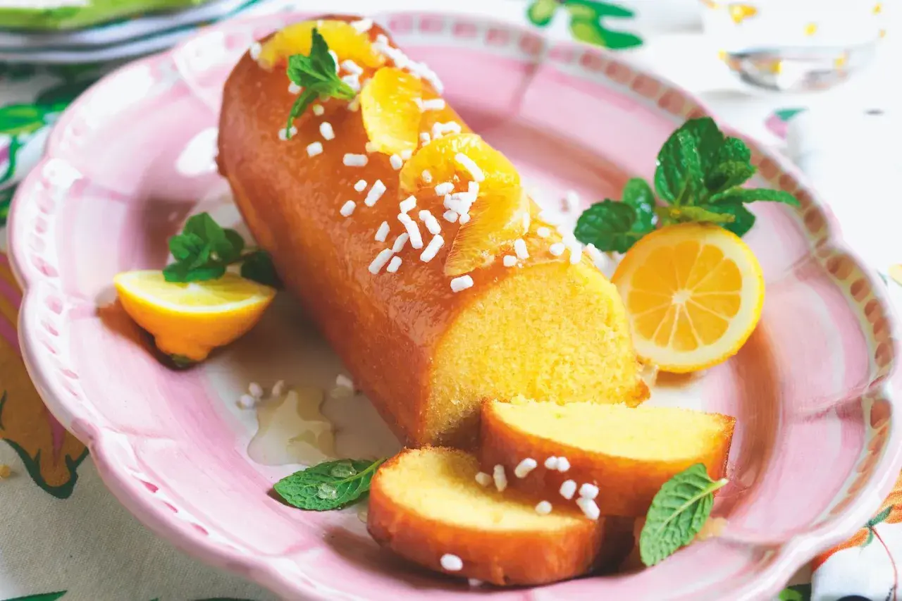 Una deliziosa torta al limone glassata, decorata con fette d'arancia, zuccherini e menta, servita su un piatto rosa con spicchi di limone.