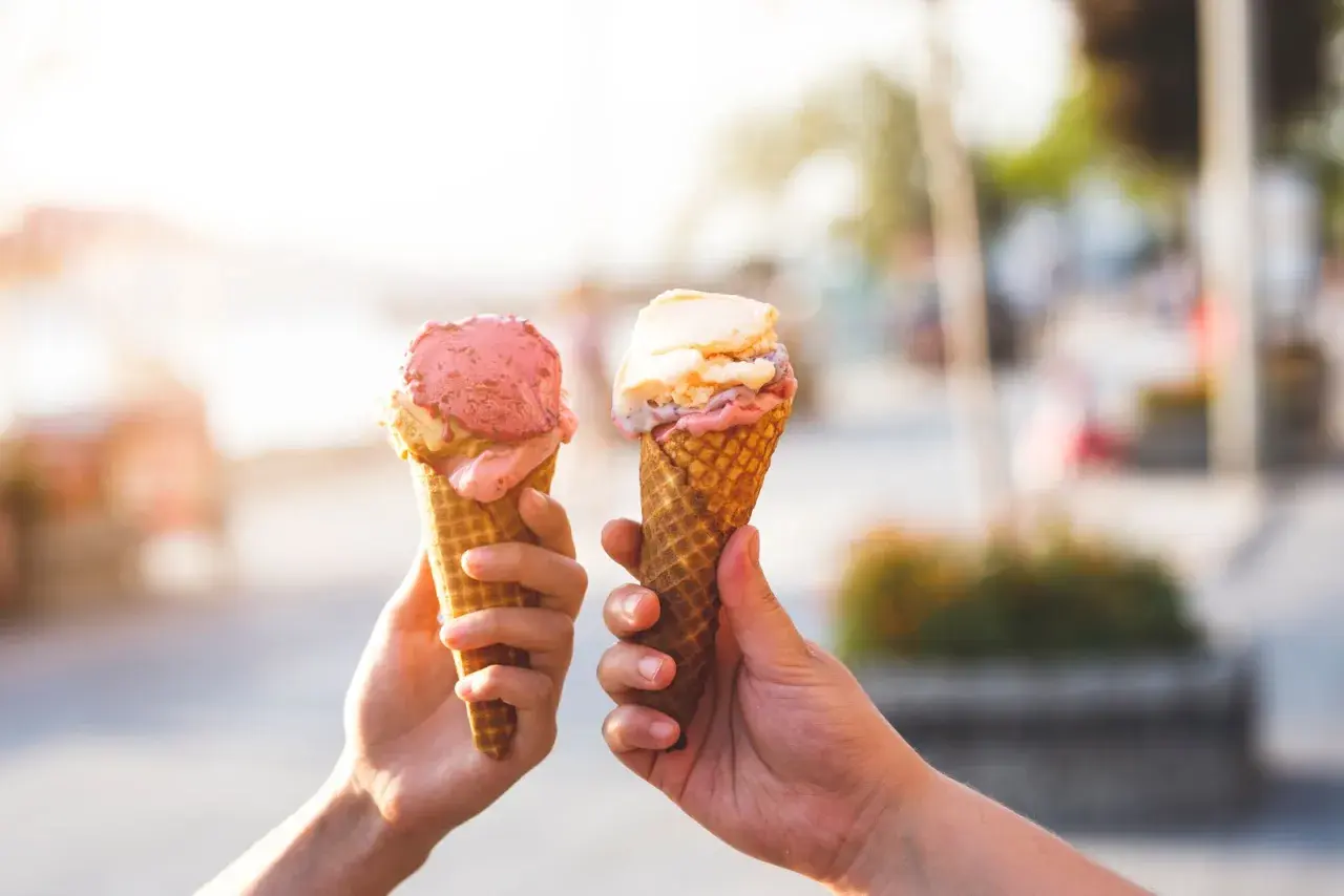 Due mani sollevano due coni gelato, uno rosa e uno con sfumature crema e rosa, celebrando la giornata del gelato.