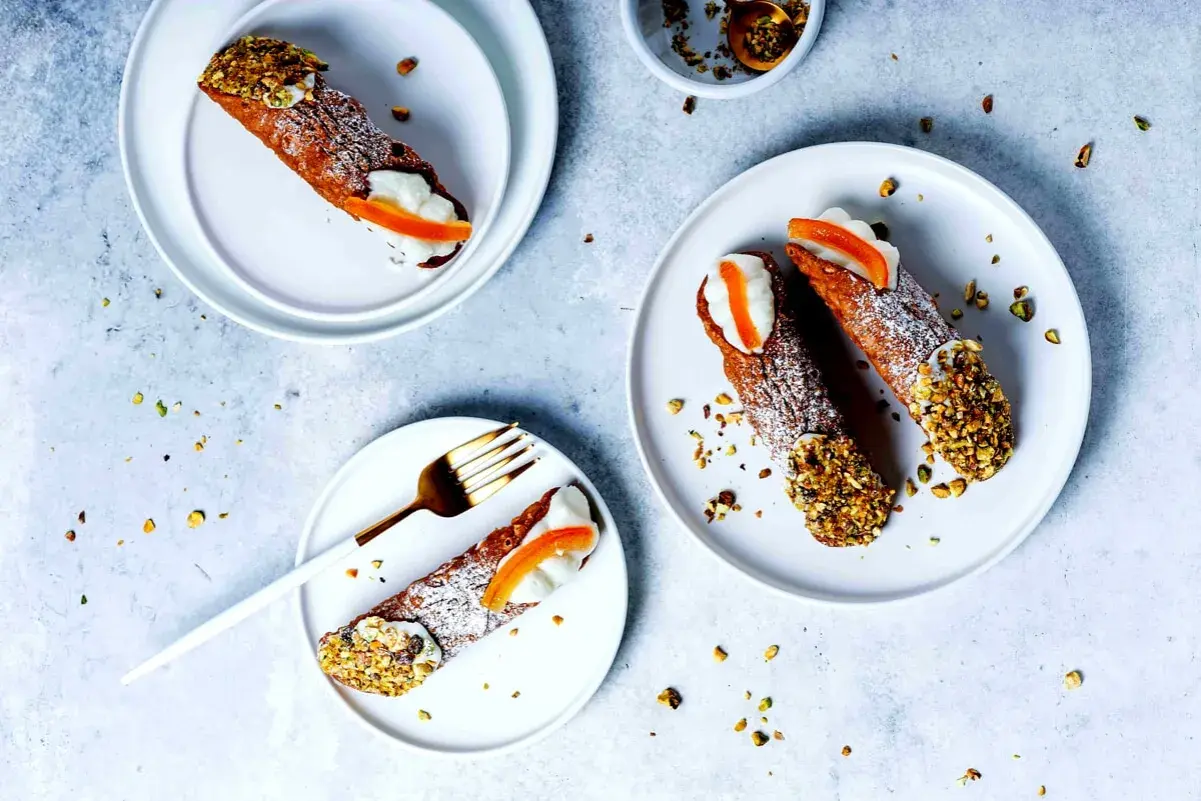 Tre cannoli siciliani, guarniti con crema, arancia candita e pistacchi tritati, su piatti bianchi, spolverati di zucchero a velo.