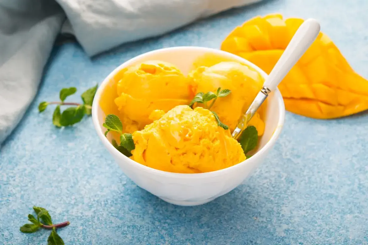 Coppa di sorbetto al mango con menta fresca e una fetta di mango sullo sfondo.