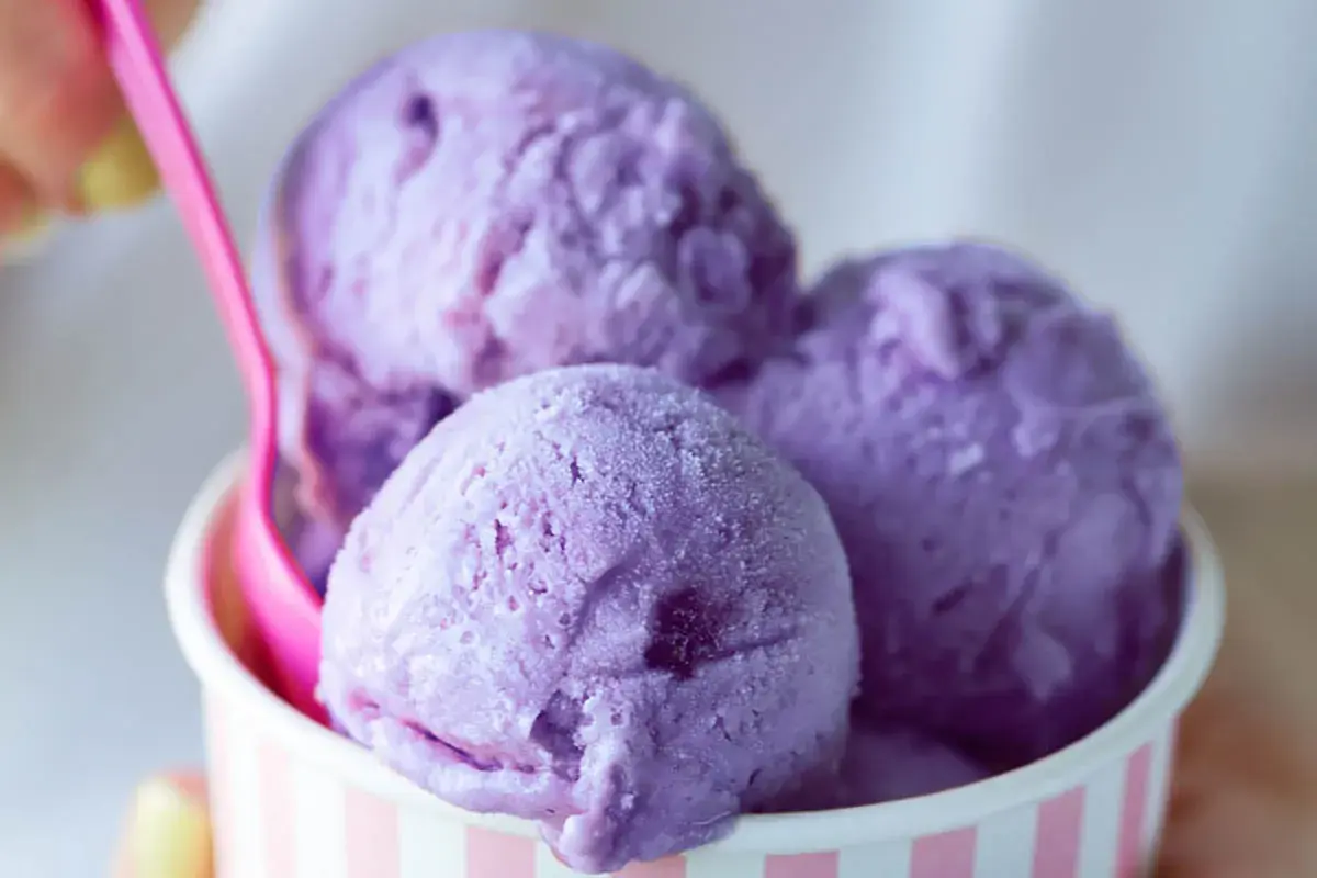 Tre palline di gelato viola in una coppetta a righe rosa e bianche, con un cucchiaino rosa. Un dolce piacere che non causa stitichezza.