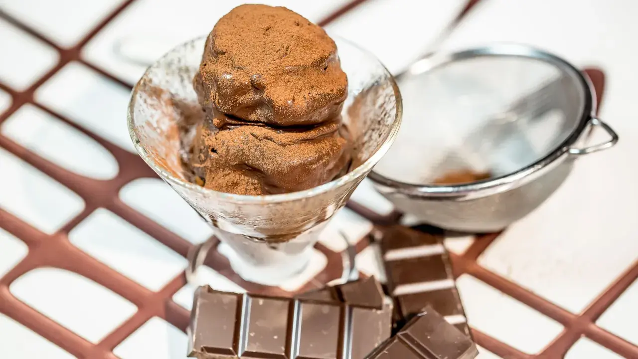 Coppetta di gelato al cioccolato spolverata di cacao, con tavolette di cioccolato e colino sullo sfondo.