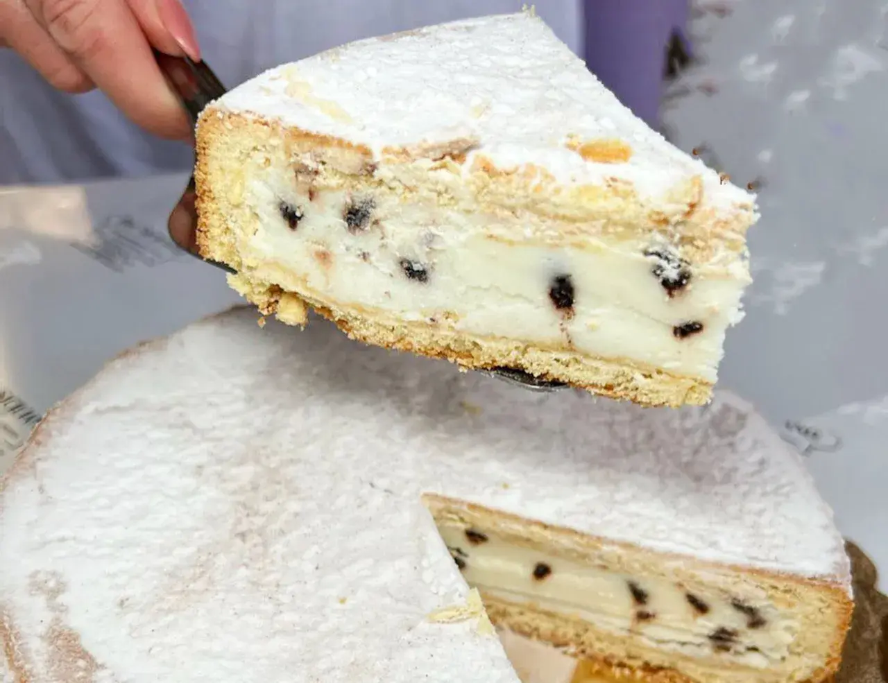 Una fetta di cassata al forno, ricca di crema e gocce di cioccolato, viene sollevata da una torta intera cosparsa di zucchero a velo.