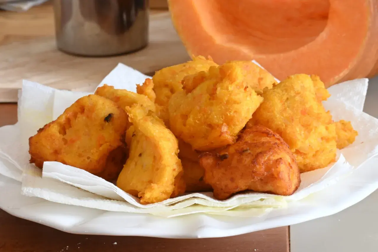 Frittelle di zucca dorate e croccanti, servite su carta assorbente, con una fetta di zucca sullo sfondo.