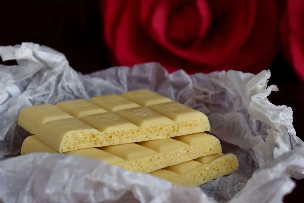 Barrette di cioccolato bianco, con la loro texture ariosa, avvolte in carta crespa, sullo sfondo di una rosa rossa. Ecco com'è fatto il cioccolato bianco.