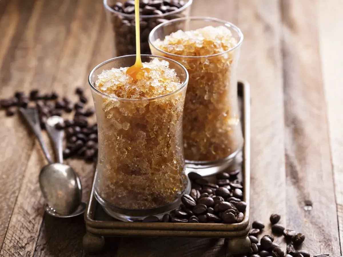 Due bicchieri di granita al caffè, con sciroppo che cola su uno. Chicchi di caffè tostati e cucchiaini completano la scena.