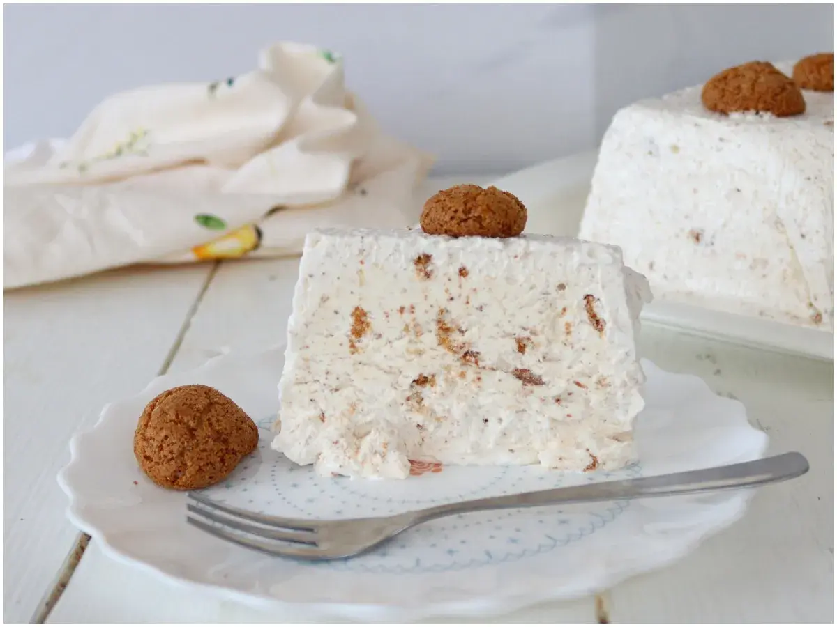 Una fetta di semifreddo agli amaretti, guarnita con un amaretto intero e un altro sul dolce intero sullo sfondo.