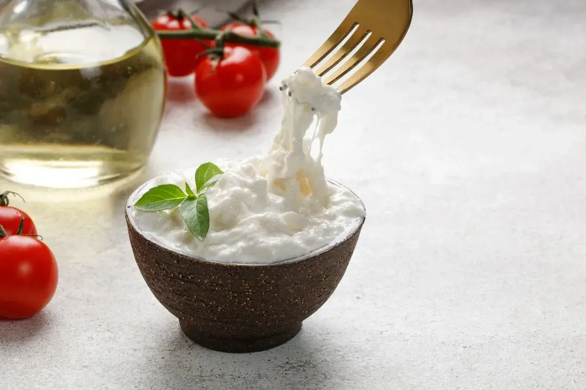 Una ciotola di stracciatella fatta in casa, con pomodorini e olio d'oliva sullo sfondo. Una forchetta solleva fili di formaggio fresco.