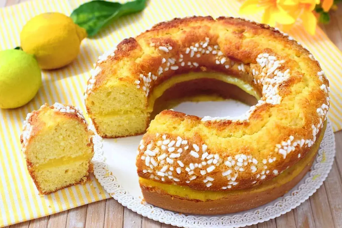 Ciambellone al limone soffice e profumato, con glassa gialla e zuccherini bianchi. Due limoni freschi e una foglia verde completano la scena.