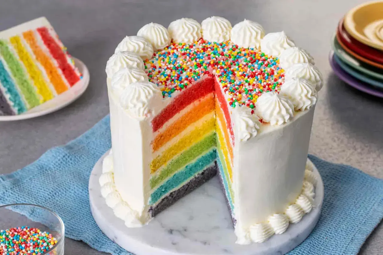 Torta arcobaleno con strati colorati, glassa bianca e zuccherini. Una fetta mostra i colori vivaci, come un vero arcobaleno.