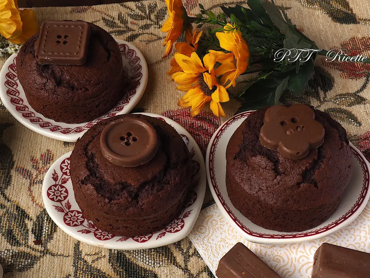 Tre deliziosi cupcake al cioccolato, decorati con bottoni di cioccolato, su piattini con fiori gialli sullo sfondo.