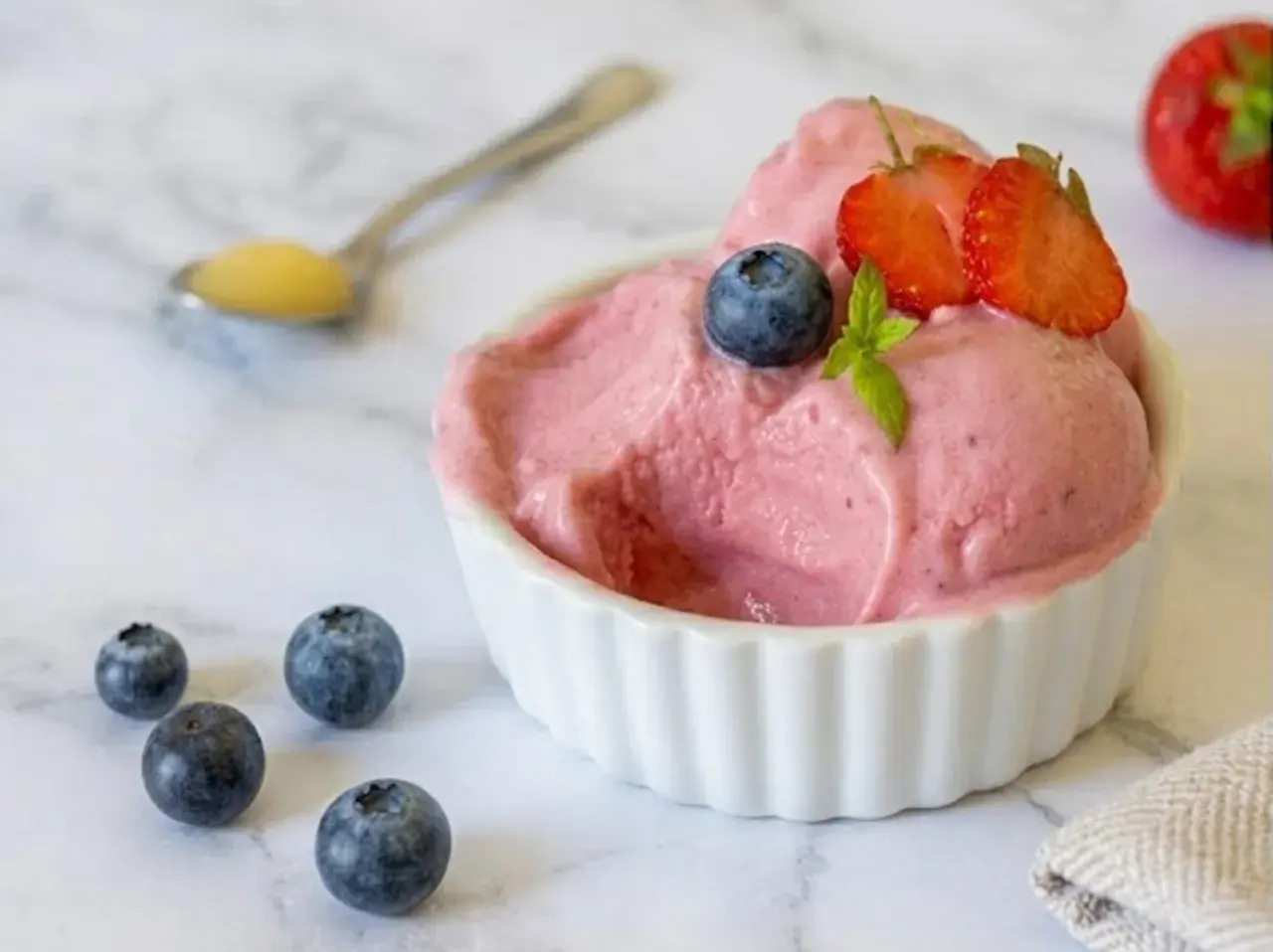 Delizioso gelato rosa, guarnito con mirtilli e fragole, servito in una ciotola bianca. Intorno, altri mirtilli e un cucchiaino con un po' di miele. Perfetto per un momento di fresco piacere, questo gelato palline di ghiaccio è un sogno.