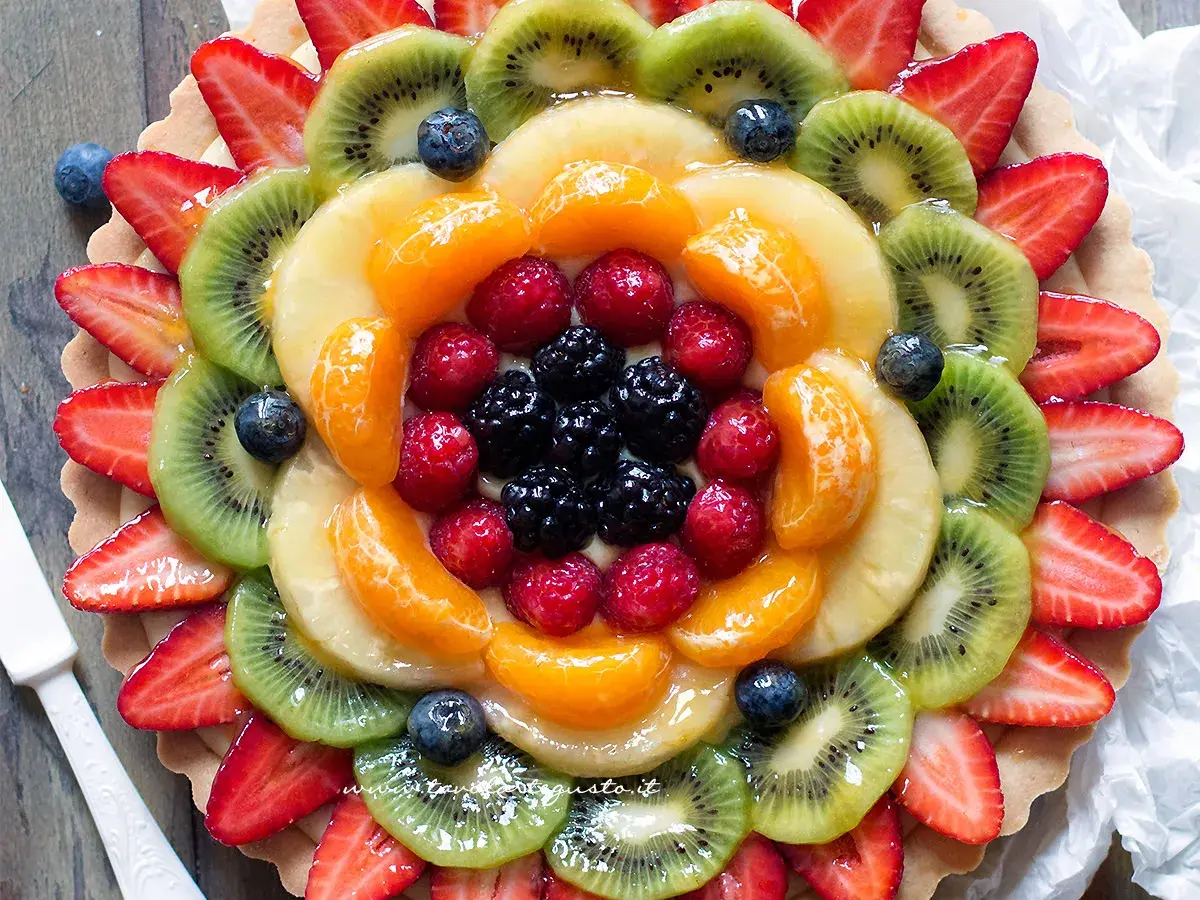 Torta alla frutta coloratissima, con fragole, kiwi, mandarini, lamponi e mirtilli disposti a raggiera su una base croccante.