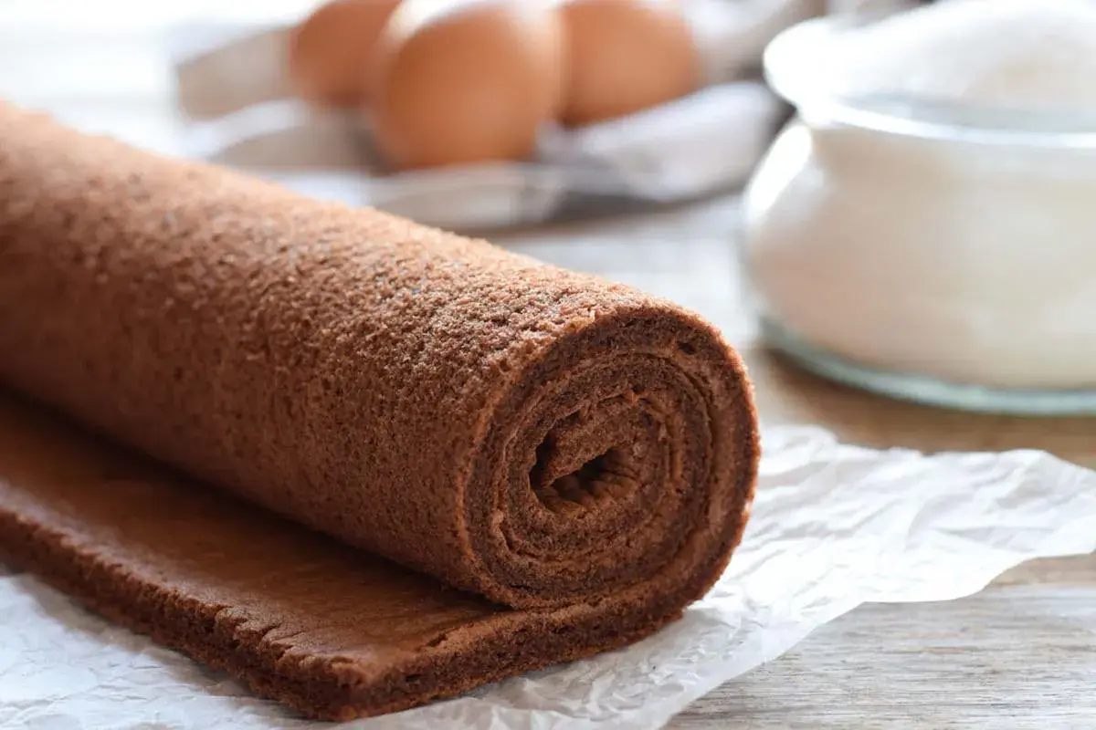 Rotolo di pasta biscotto al cacao, pronto per essere farcito. Ingredienti freschi come uova e zucchero sullo sfondo.