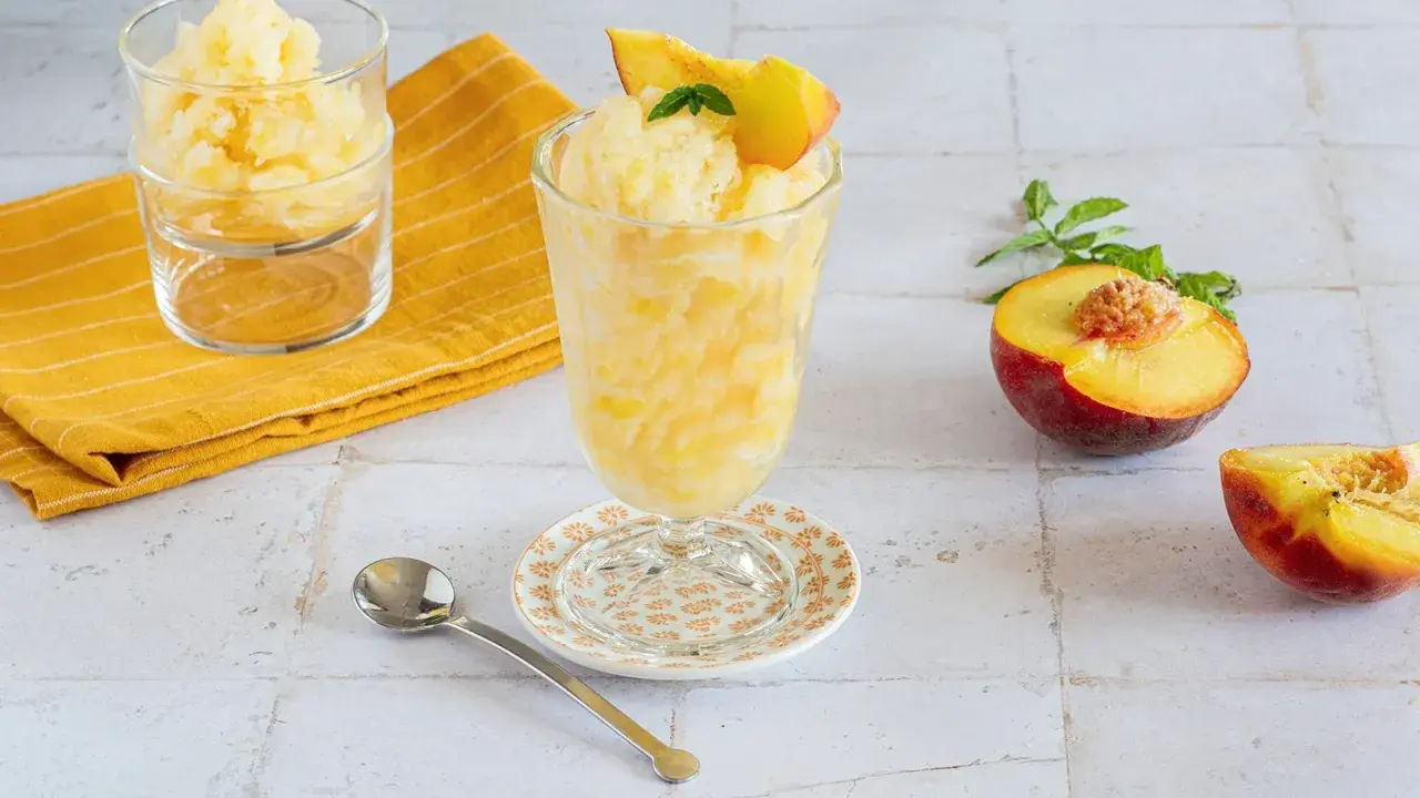 Rinfrescante granita alla pesca, guarnita con una fetta di pesca e menta. Accanto, pesche fresche e un cucchiaino.