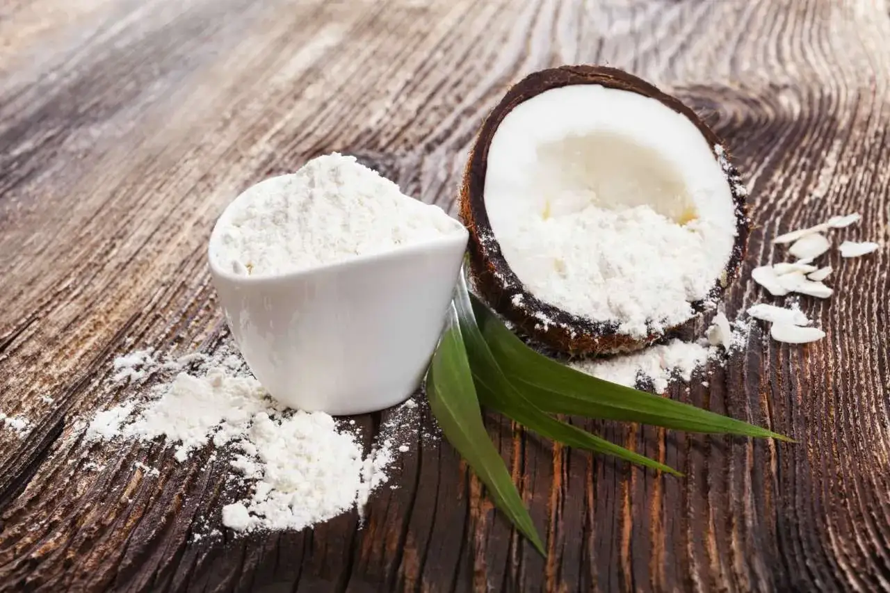 Cocco grattugiato in una ciotola bianca e mezzo cocco su un tavolo di legno. Un'ottima alternativa alla farina di cocco per le tue ricette.