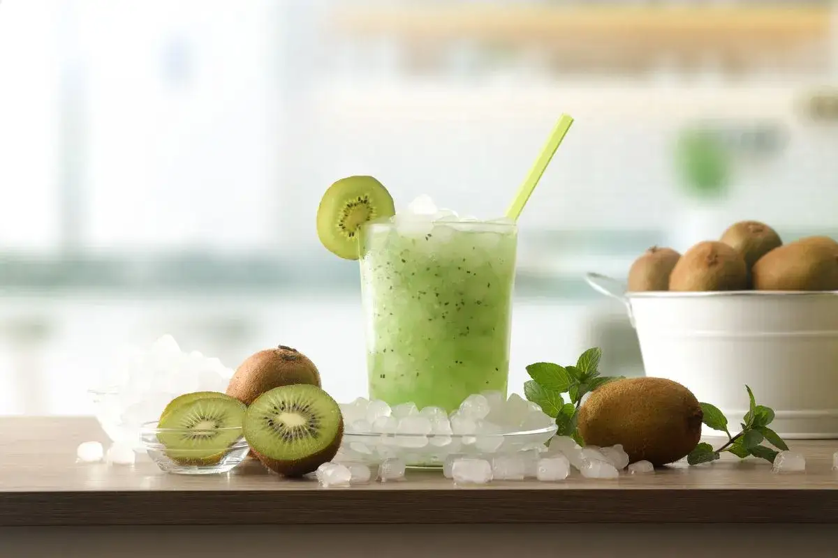 Un rinfrescante sorbetto al kiwi, guarnito con una fetta di frutto e una cannuccia verde, circondato da kiwi freschi e cubetti di ghiaccio.