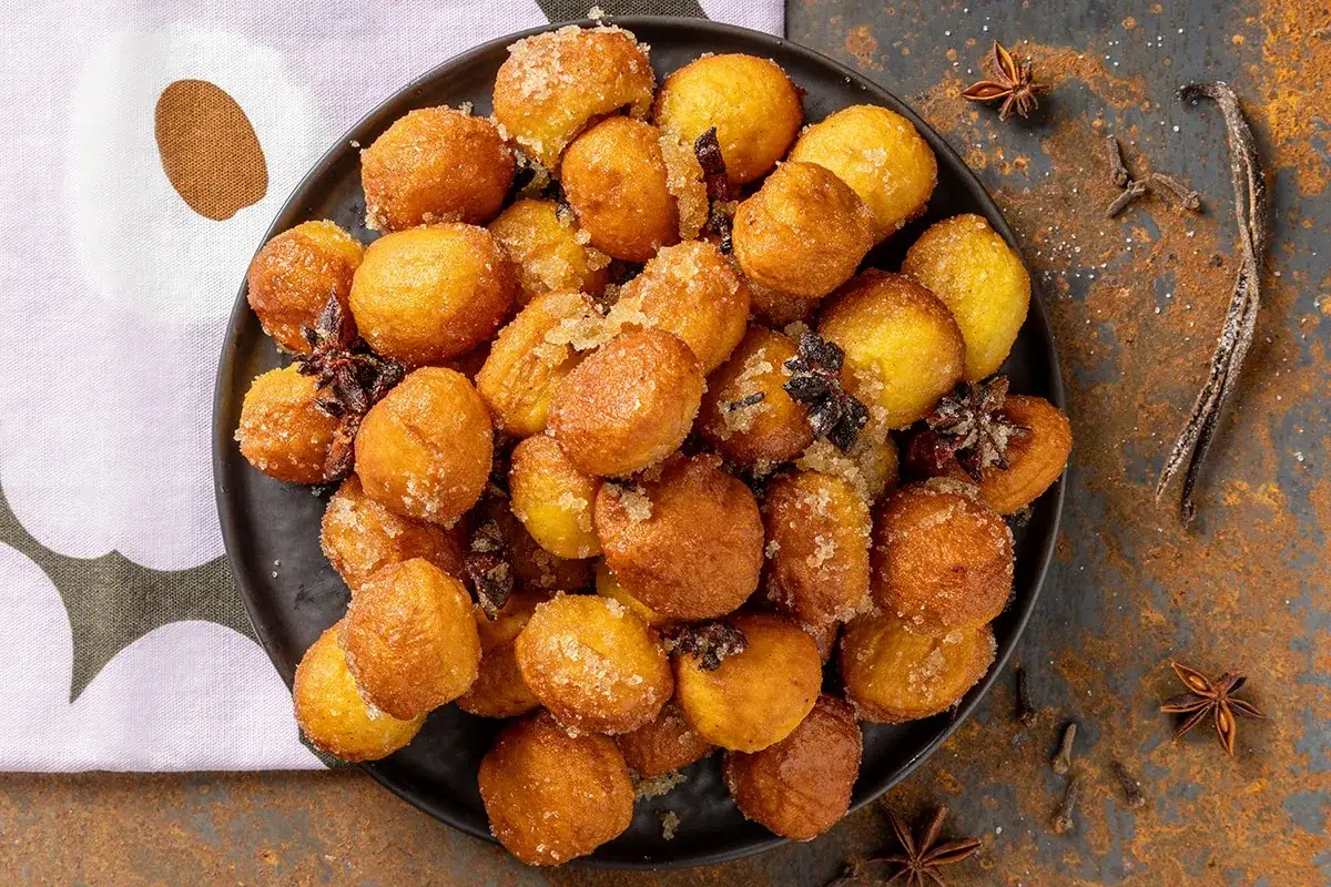 Piatto di frittelle di zucca dorate, cosparse di zucchero e spezie, con anice stellato e bacca di vaniglia sullo sfondo.