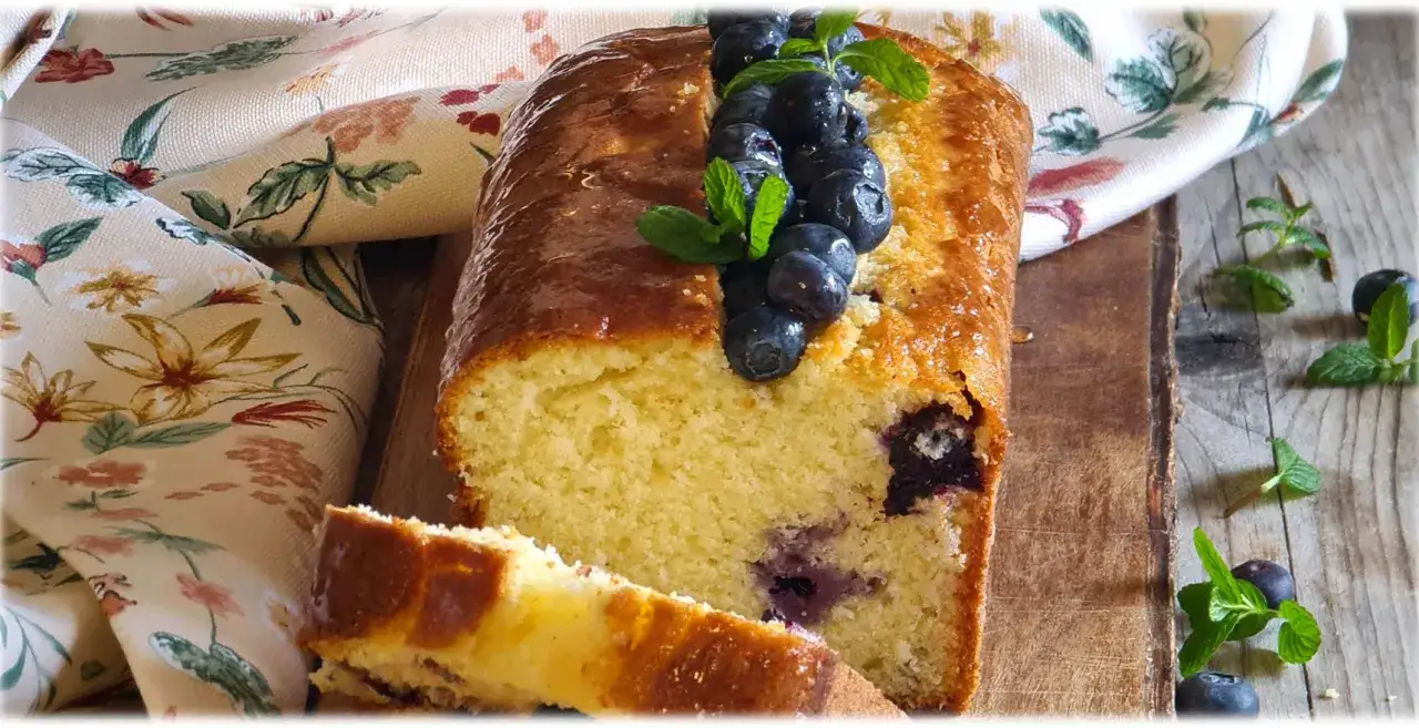 Plumcake ai mirtilli, soffice e dorato, guarnito con mirtilli freschi e foglioline di menta su un tagliere di legno.