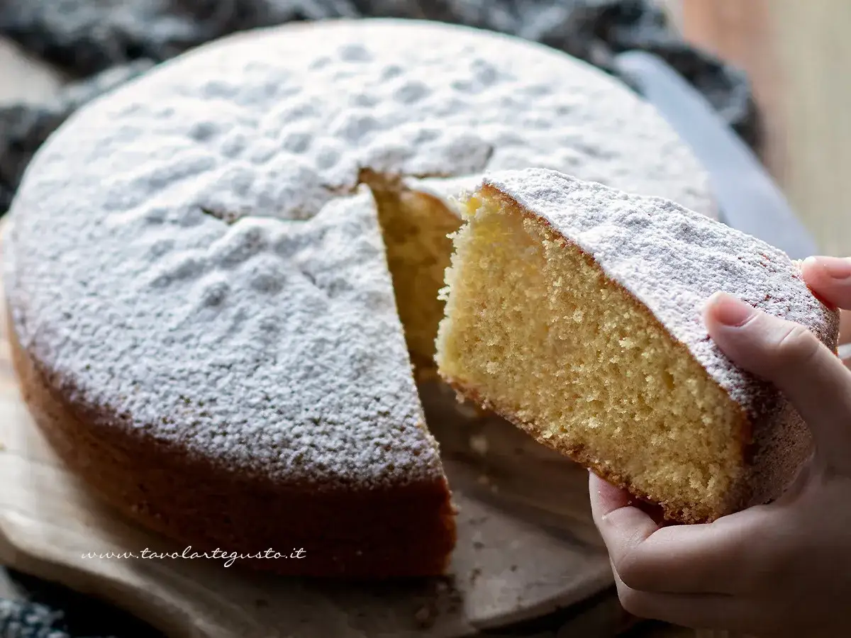 Una fetta di torta margherita soffice, cosparsa di zucchero a velo, viene sollevata da una mano.
