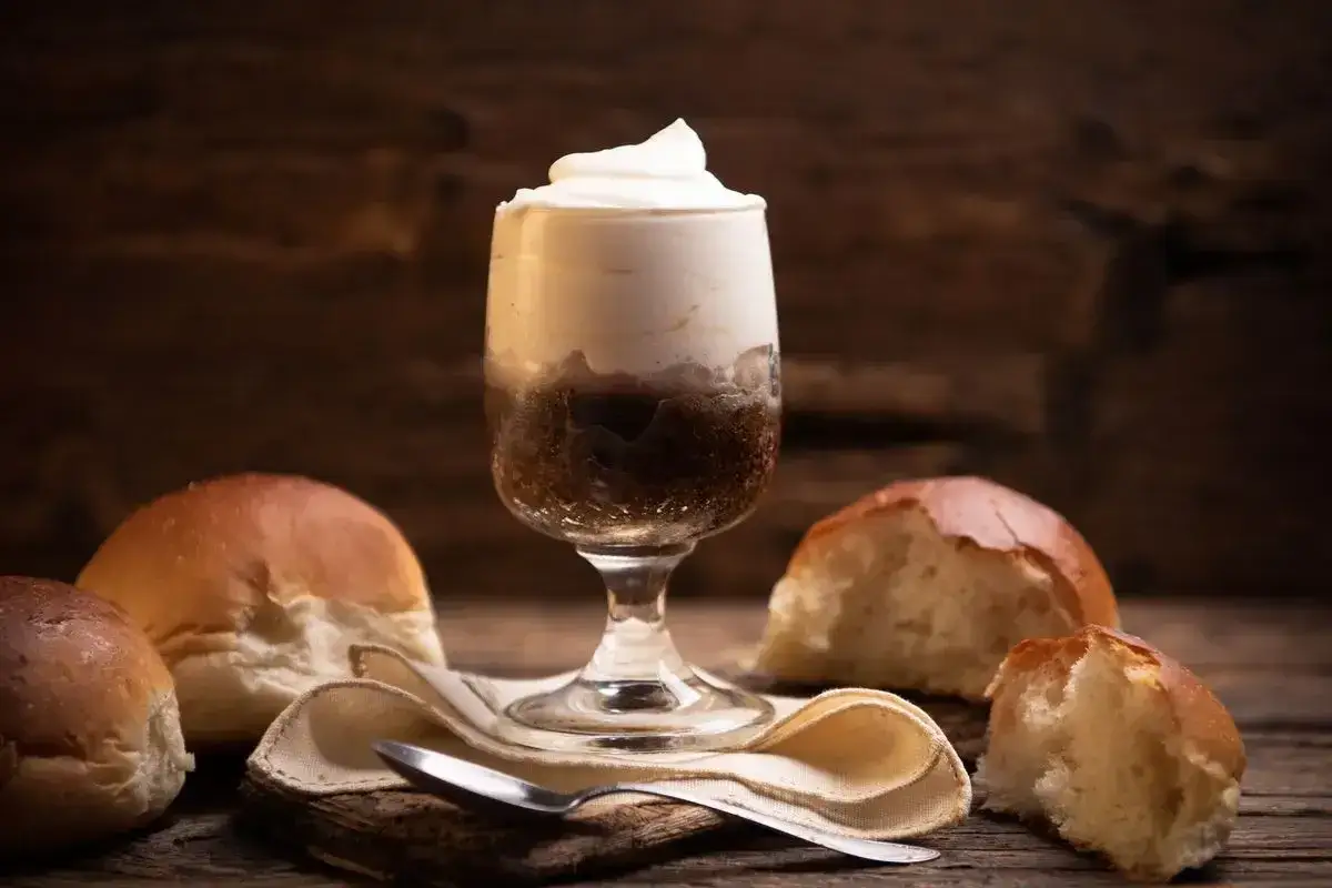 Granita al cioccolato catanese senza gelatiera, servita in un bicchiere con panna montata, accompagnata da soffici brioche su un tavolo di legno.