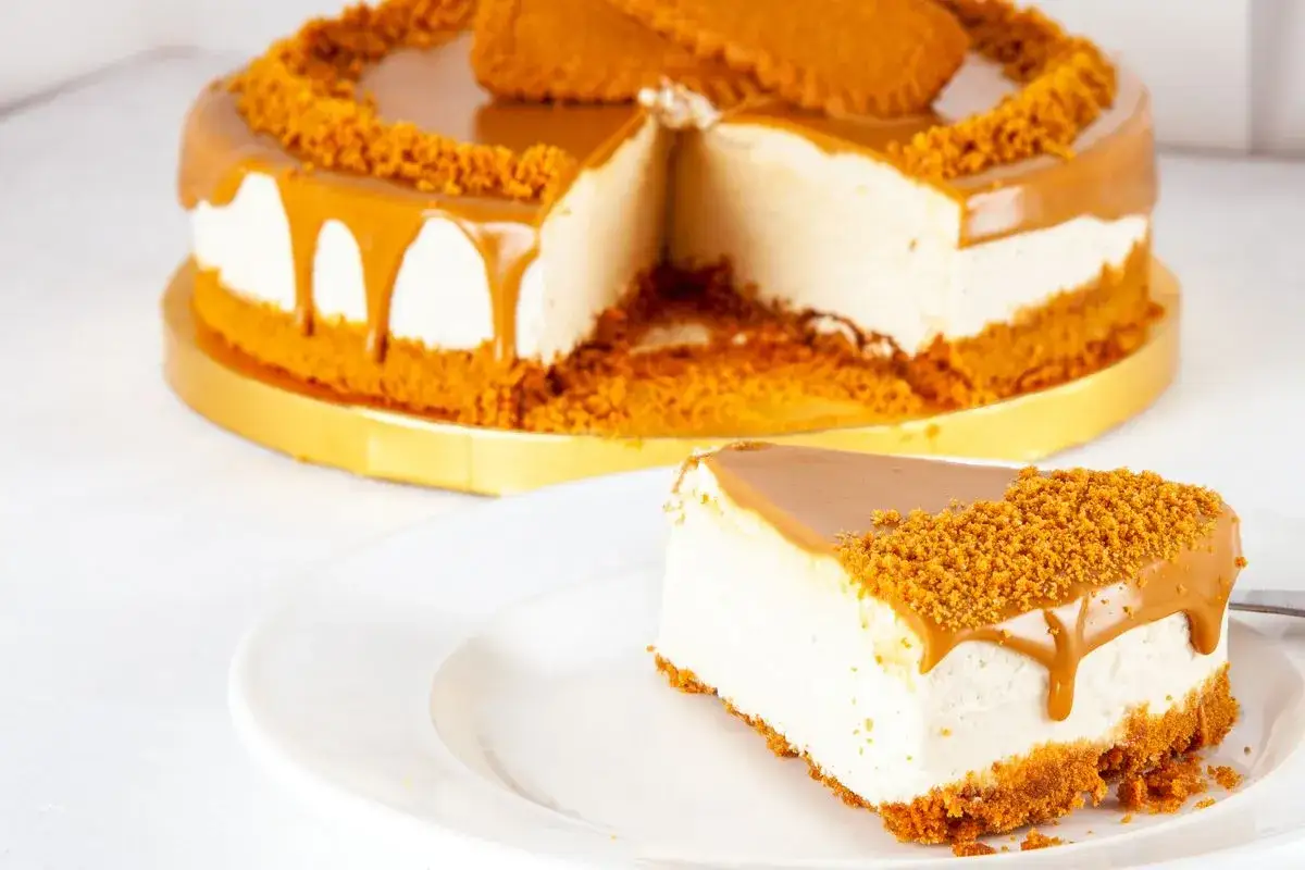 Una fetta di torta Lotus su un piatto bianco, con la torta intera decorata con biscotti e caramello sullo sfondo.