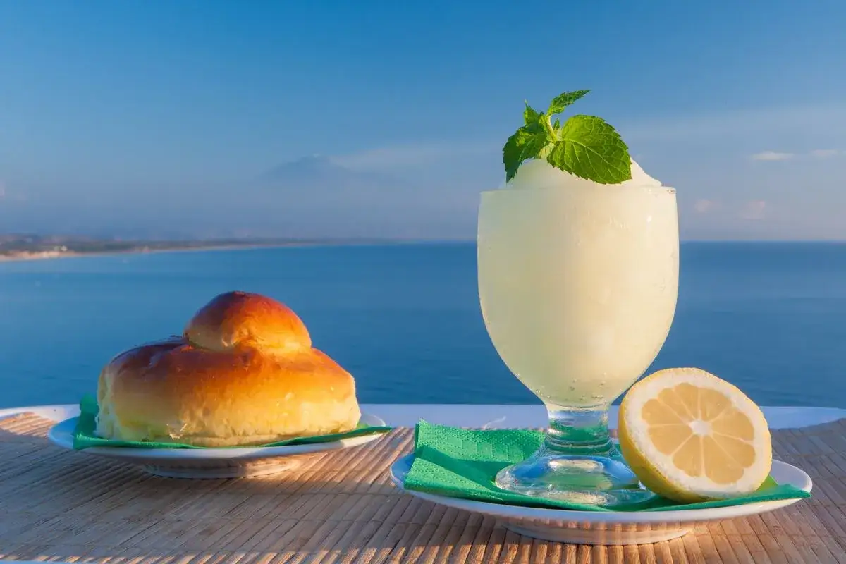 Granita al limone, brioche e limone fresco con mare e vulcano sullo sfondo. Perfetta ricetta granita al limone per una colazione siciliana.