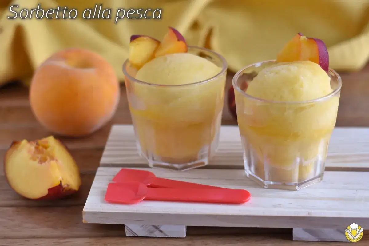 Due bicchieri di sorbetto alla pesca, guarniti con spicchi di pesca fresca. Una pesca intera e uno spicchio sono visibili sul tavolo.