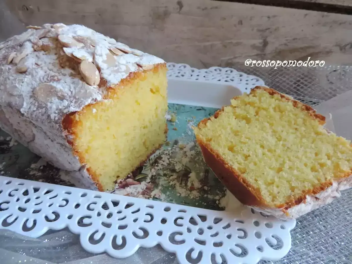Plumcake yogurt greco soffice, cosparso di zucchero a velo e mandorle a lamelle. Una fetta mostra la sua morbidezza.