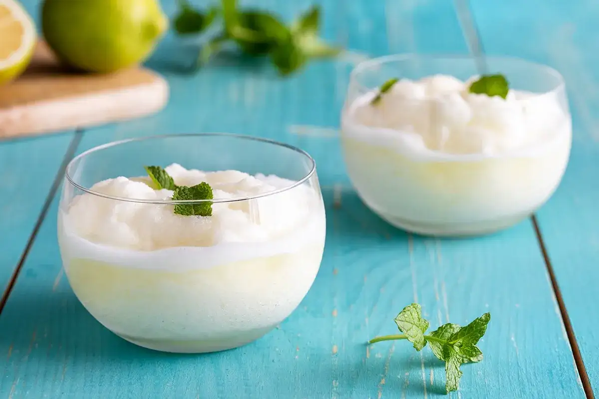Due bicchieri di sorbetto limone senza albume, guarniti con menta fresca, su un tavolo di legno blu.