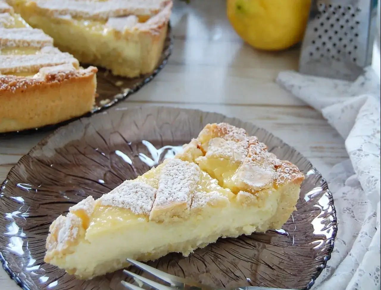 Una fetta di babbaiola posillipo, crostata con crema gialla e decorata con zucchero a velo, su un piatto di vetro scuro.