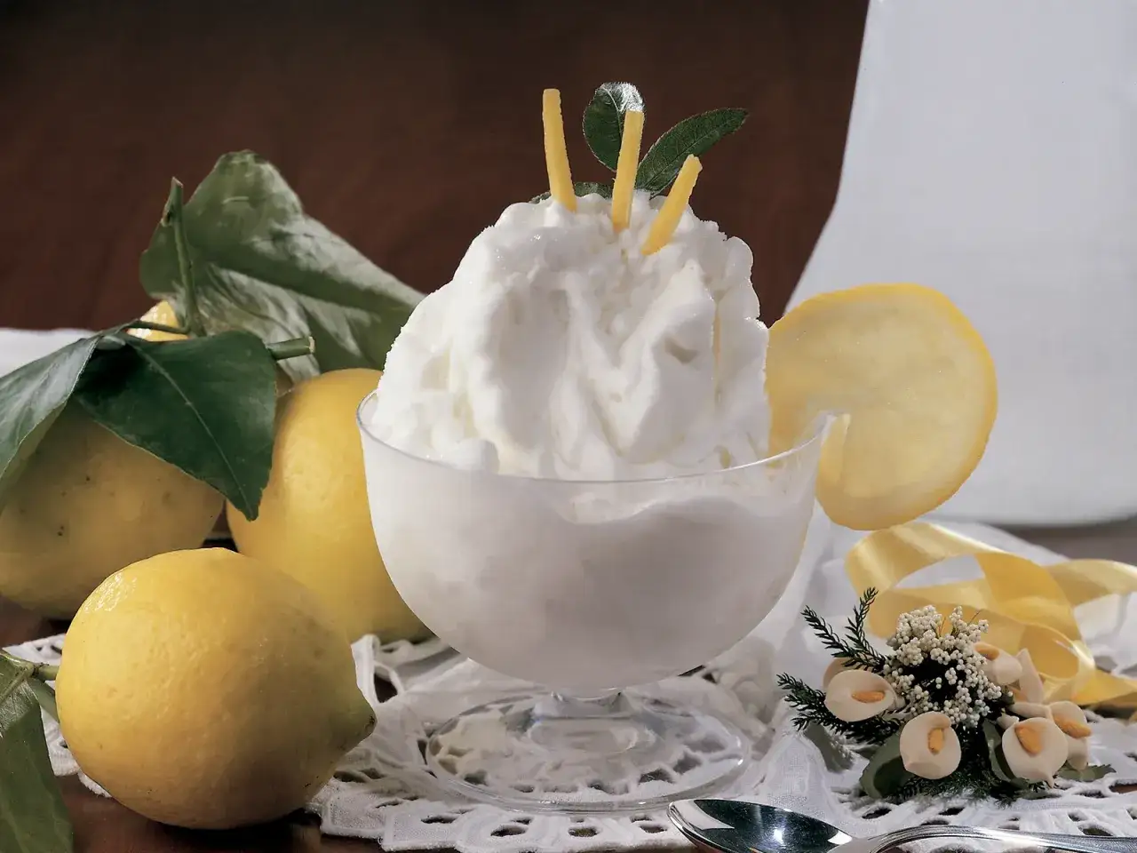 Coppa di sorbetto al limone con gelato, guarnita con scorzette e foglie, circondata da limoni freschi.