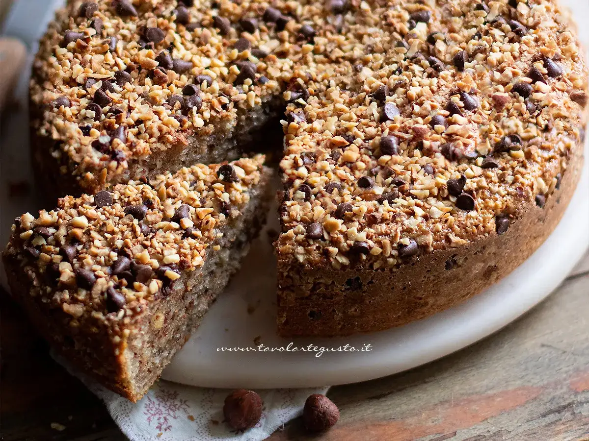 Deliziosa torta di nocciole piemontese, ricca di nocciole tritate e gocce di cioccolato, con una fetta tagliata.