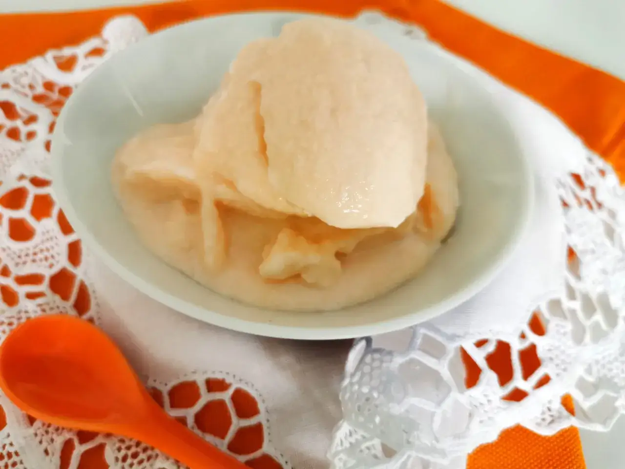 Delizioso gelato al melone solo frutta, servito in una ciotola bianca su un tovagliolo di pizzo bianco e un runner arancione. Un cucchiaio arancione è posizionato a sinistra.