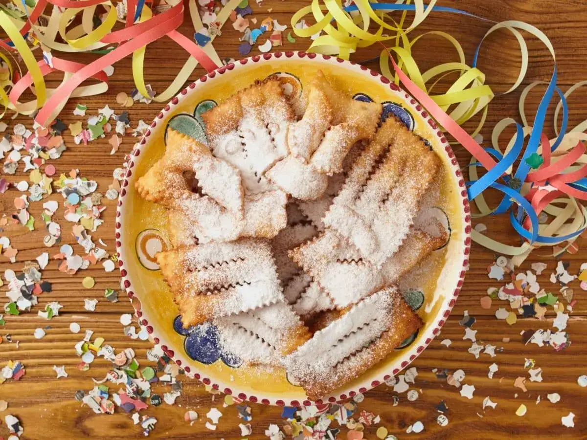 Chiacchiere di Carnevale Benedetta Rossi, cosparse di zucchero a velo, su un piatto decorato, circondate da coriandoli e stelle filanti.