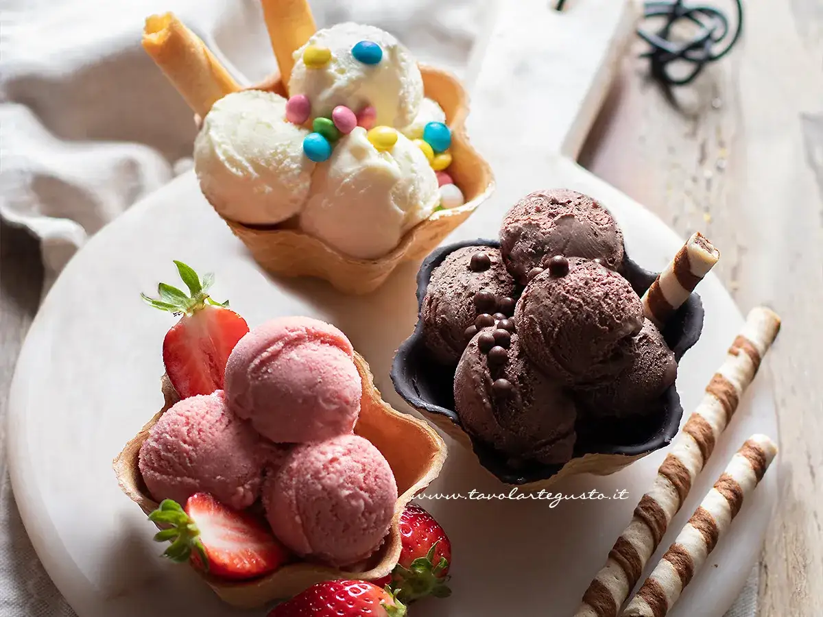 Tre coppette di gelato: fragola, cioccolato e vaniglia con palline di ghiaccio colorate e cialde.