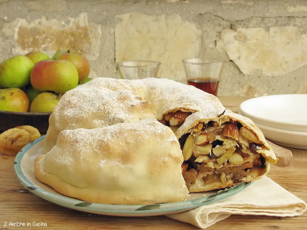 Strudel di mele e mandorle, una rocciata umbra spolverata di zucchero a velo, con mele fresche e bicchierini di liquore sullo sfondo.