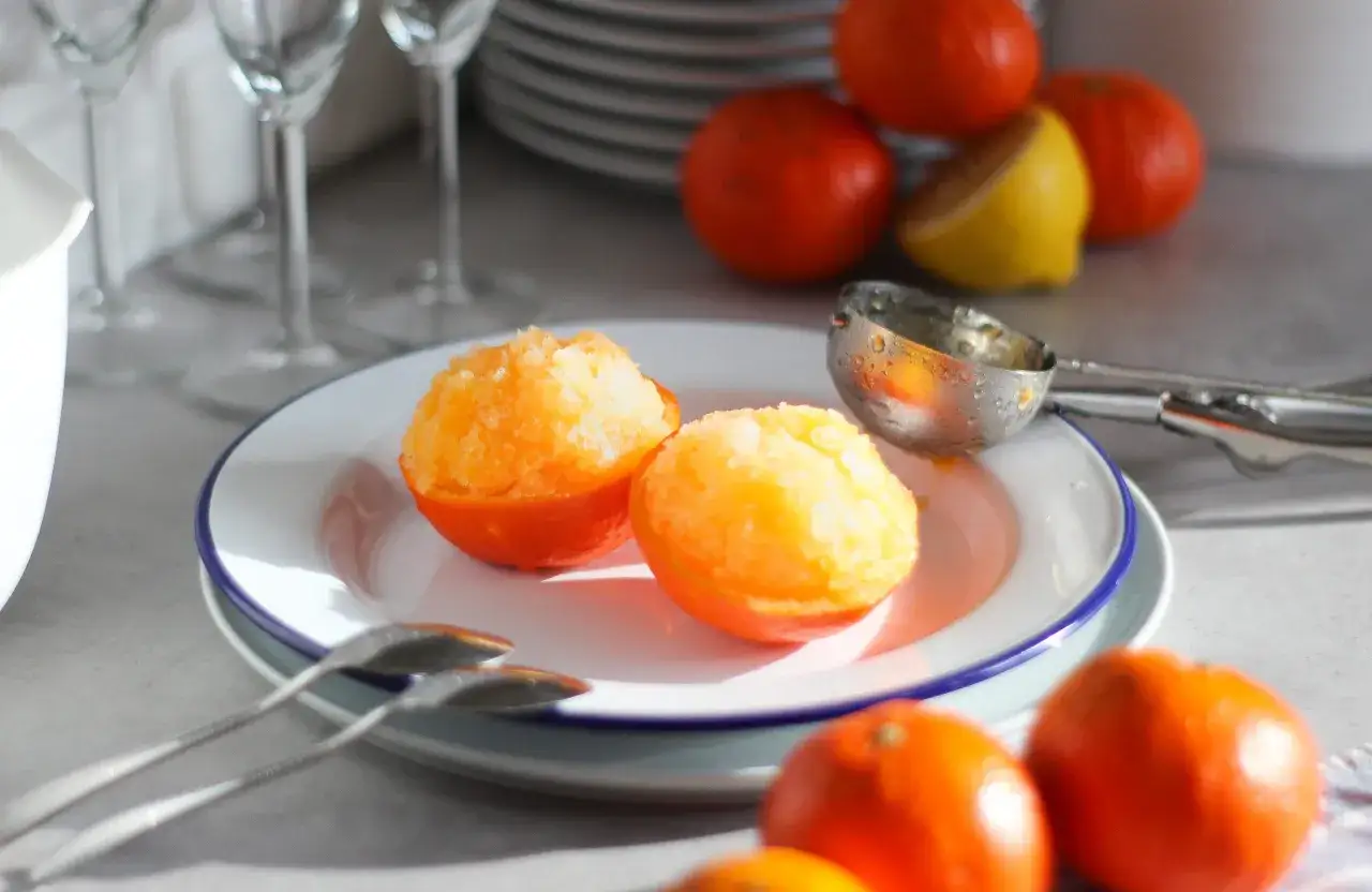 Sorbetto al mandarino senza gelatiera servito in coppette di mandarino. Frutta fresca e cucchiaio da gelato sullo sfondo.