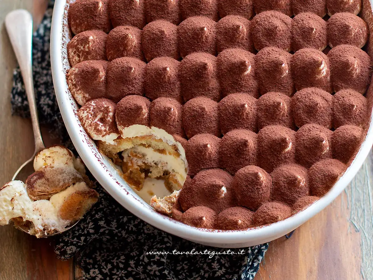 Una deliziosa torta tiramisù, ricoperta di cacao amaro, pronta per essere gustata. Un cucchiaio ne ha già prelevato una porzione.