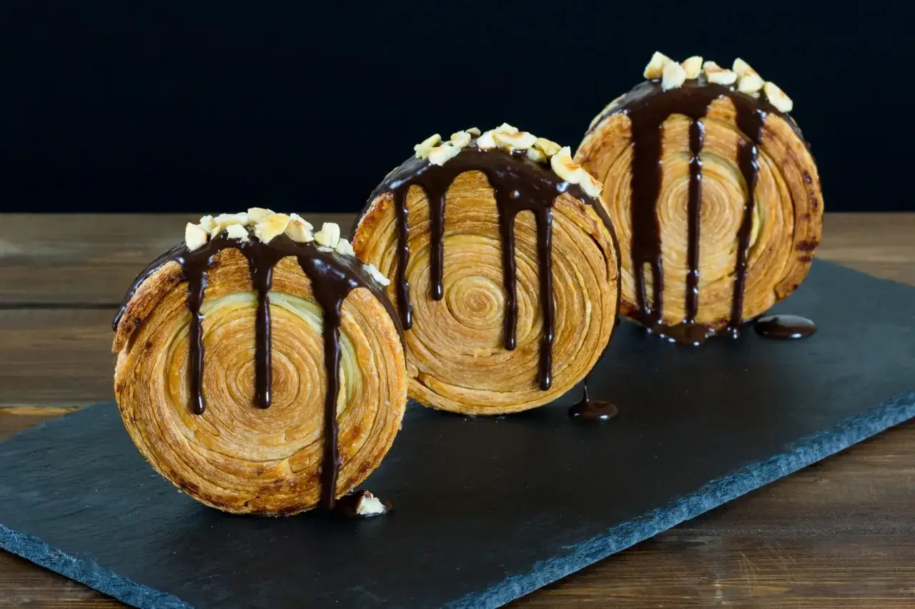 Tre deliziosi new york roll, ricoperti di cioccolato fuso e granella di nocciole, su una lavagna scura.