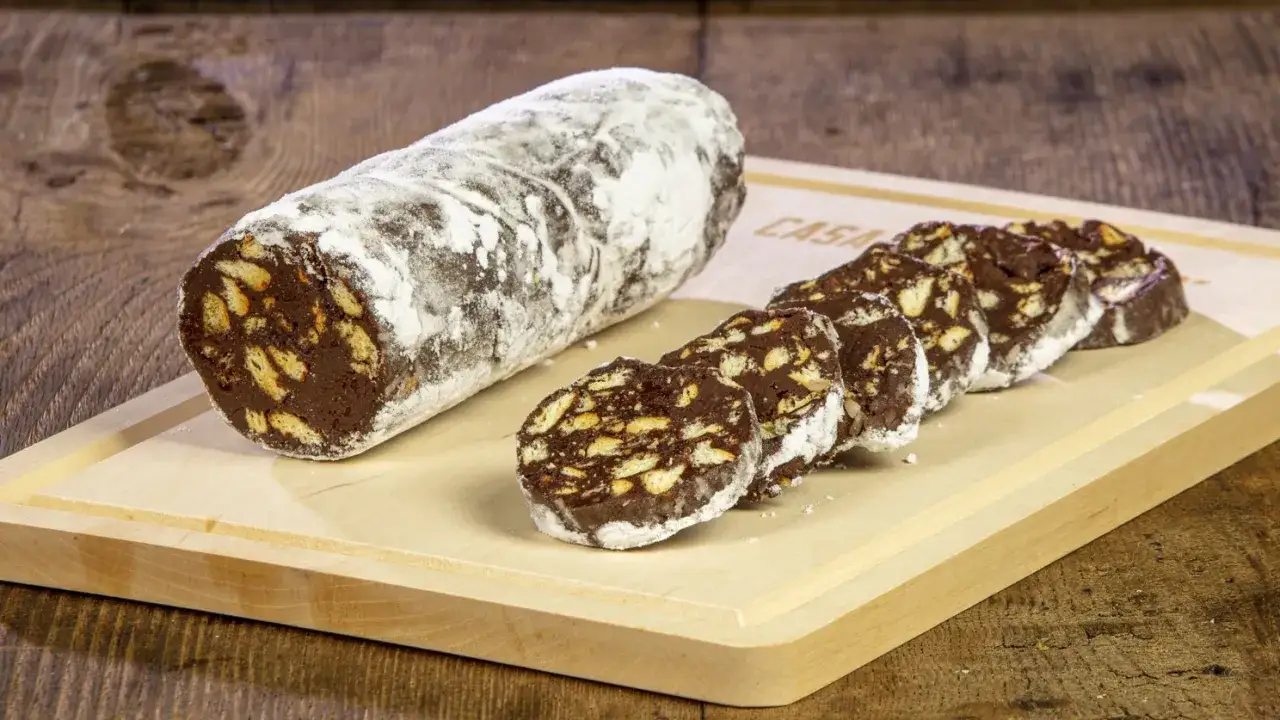 Un rotolo di salame al cioccolato, cosparso di zucchero a velo, con fette tagliate su un tagliere di legno.