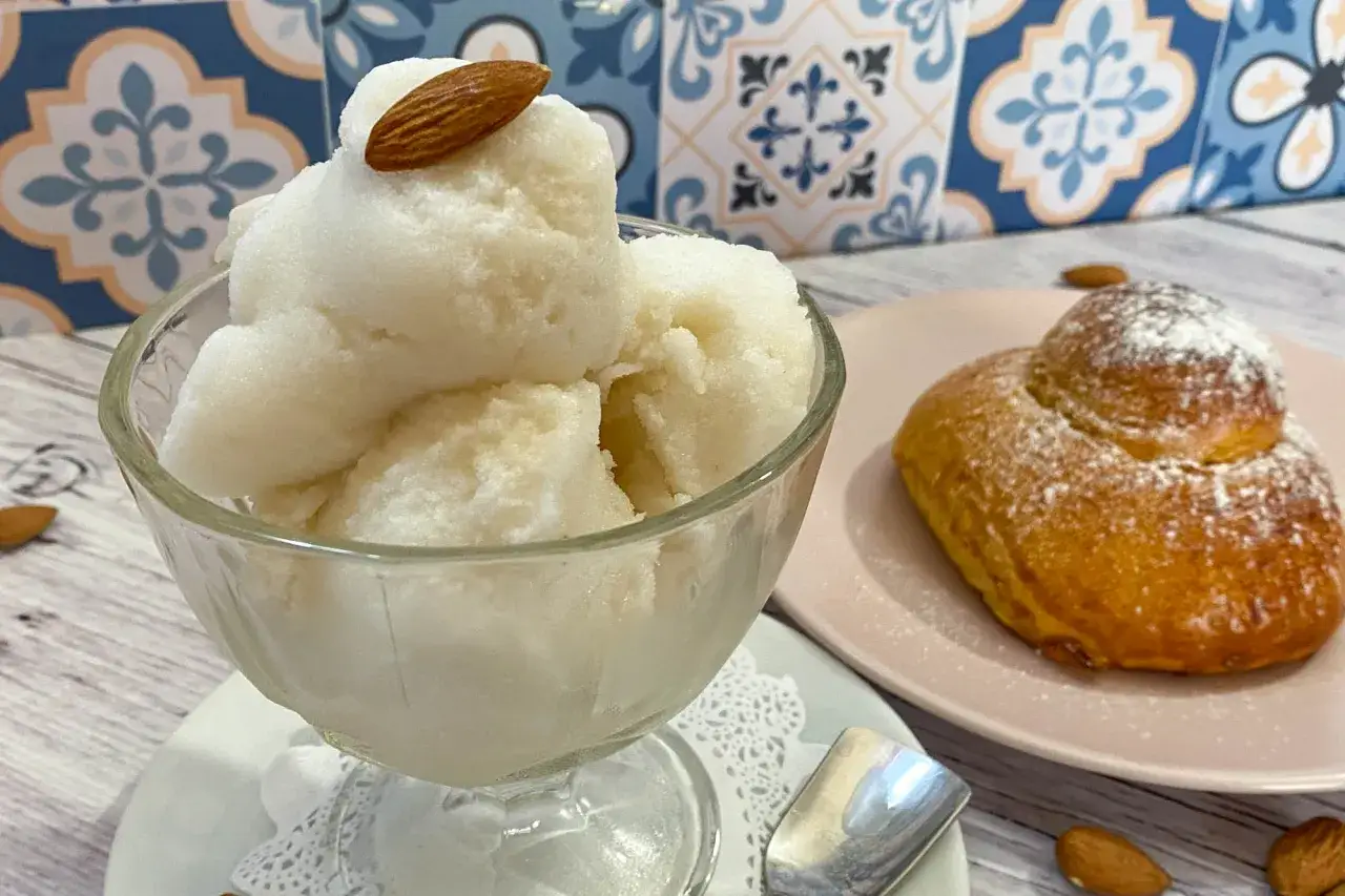 Coppa di granita di mandorle, guarnita con una mandorla intera, accanto a un brioche spolverato di zucchero a velo.