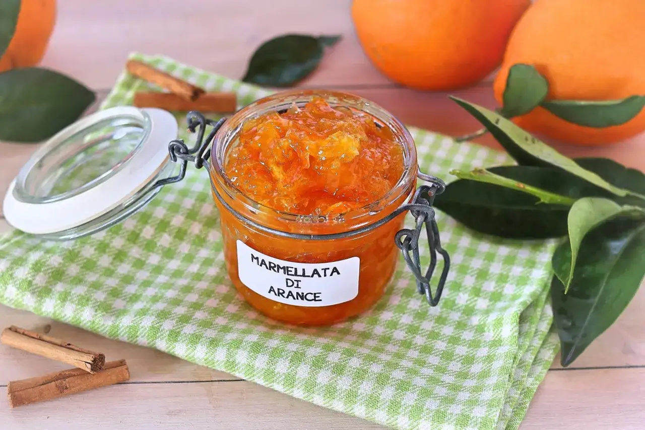 Un vasetto di marmellata di arance fatto in casa, con arance fresche e bastoncini di cannella sullo sfondo.