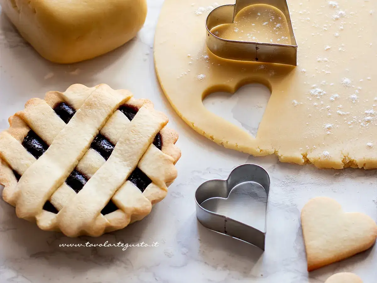 Preparazione di crostatine con pasta frolla: impasto, stampini a cuore e una crostatina gi&agrave; pronta con ripieno di marmellata.
