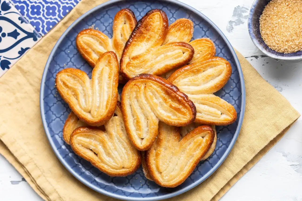 Un piatto blu pieno di dolci con pasta sfoglia dorati e croccanti, cosparsi di zucchero.